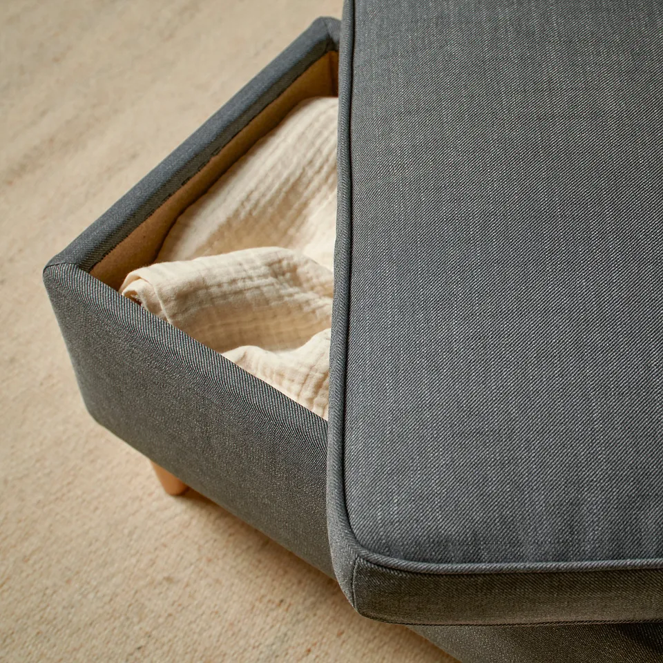 A grey fabric footstool with a lid, providing storage space underneath. Used for resting feet or extra seating.