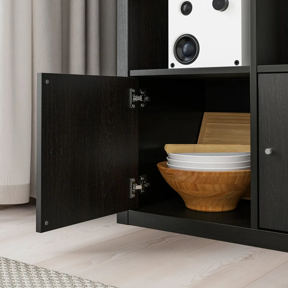 Black KALLAX storage unit with wooden bowl and plates, closed door, mounted on light wooden floor.