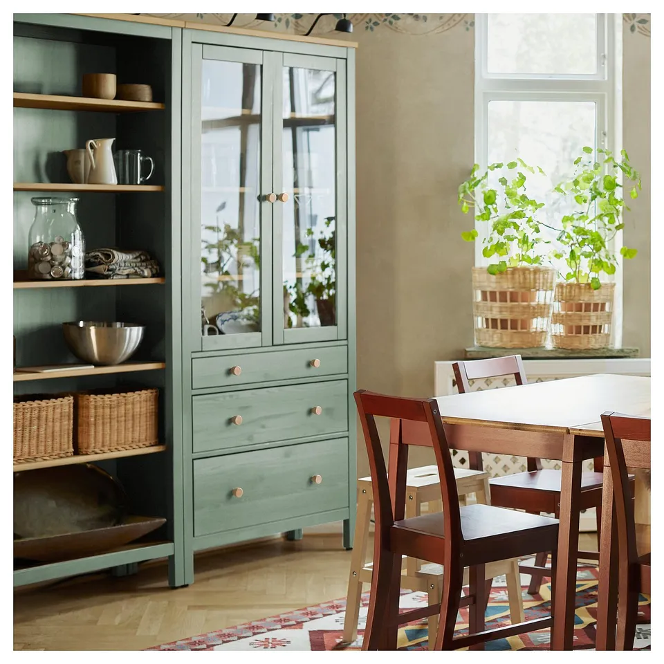 A cosy dining room a green HEMNES cabinet, wooden chairs, plants, and a colourful rug.