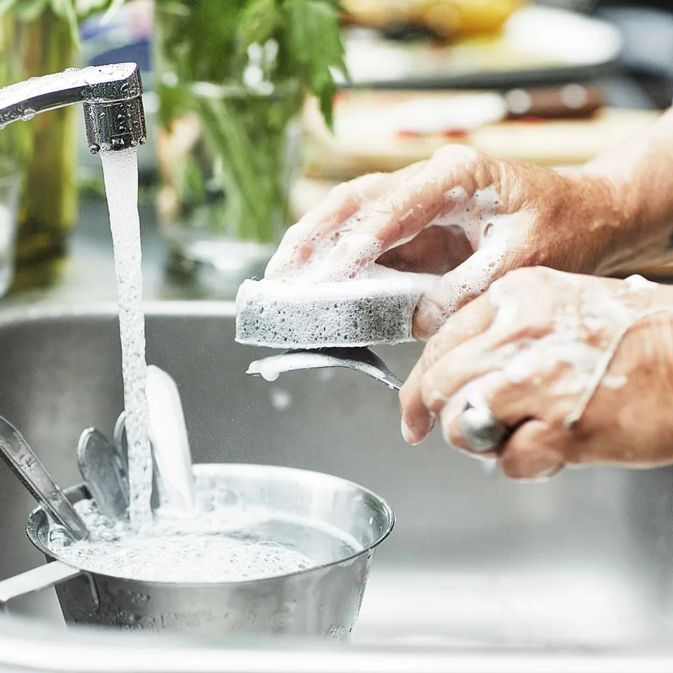A person washing dishes in a kitchen sink with a KALLSJÖN chrome-plated brass tap, featuring a swivel spout.
