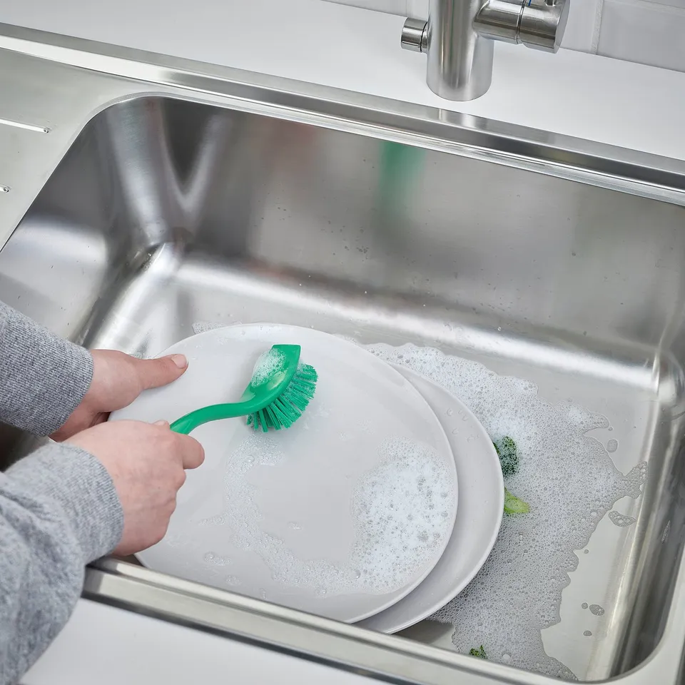 Person scrubbing dish with green brush in stainless steel sink.