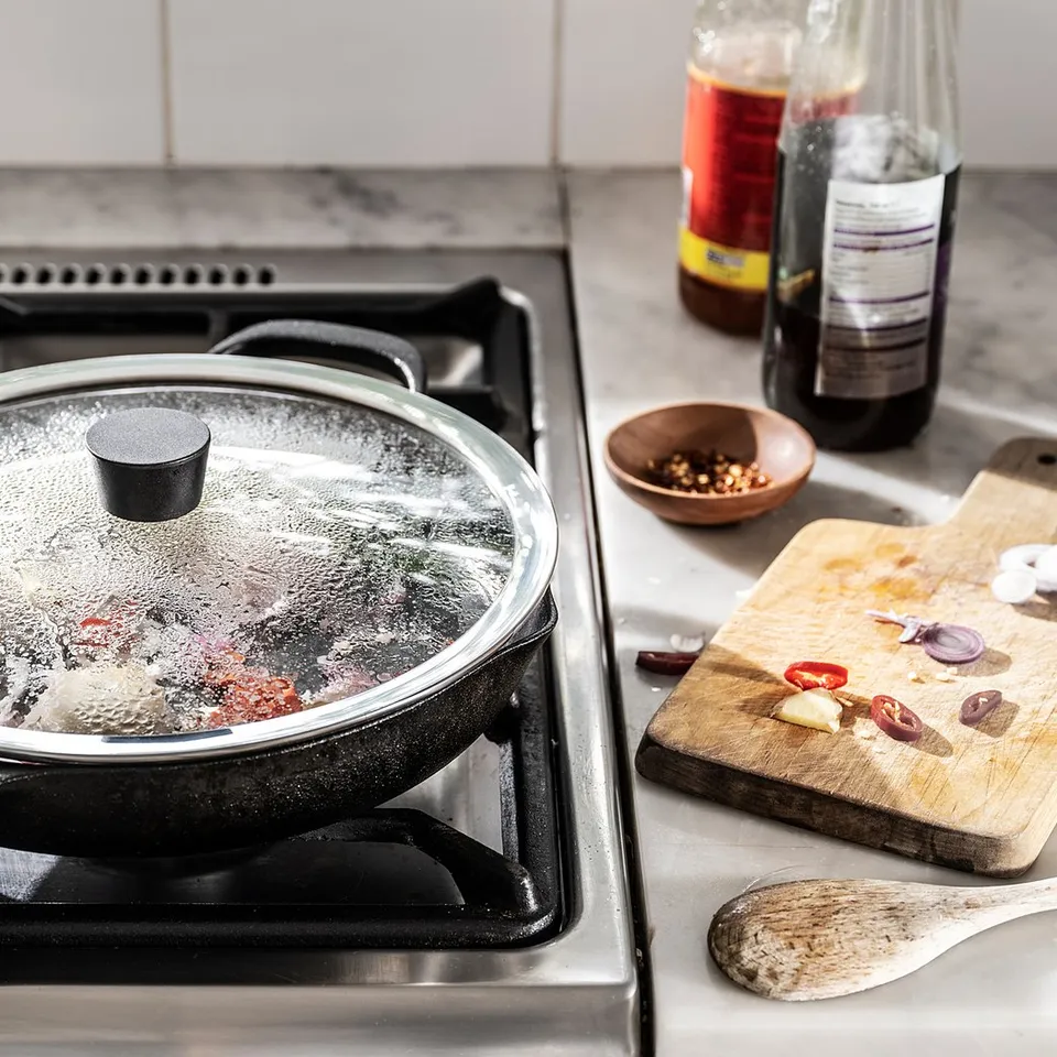 A covered frying pan on a stove. Nearby, two bottles and chopped vegetables on a wooden board.