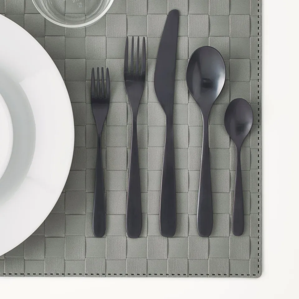 Modern black cutlery set on grey place mat, including fork, knife, and spoon, shown with white plate and glass.