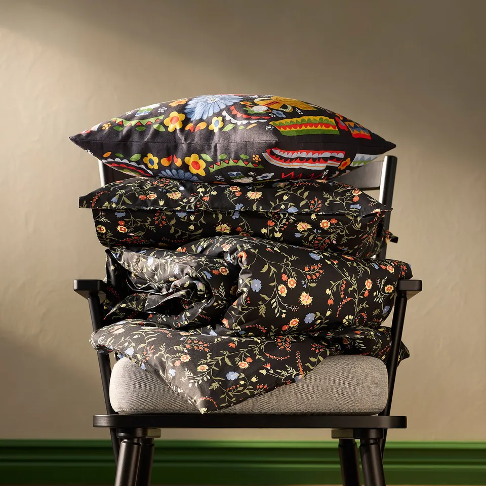 Black BRUDKRUSBÄR pillows on chair. Floral pattern, stacked in various sizes. Soft, organic cotton.