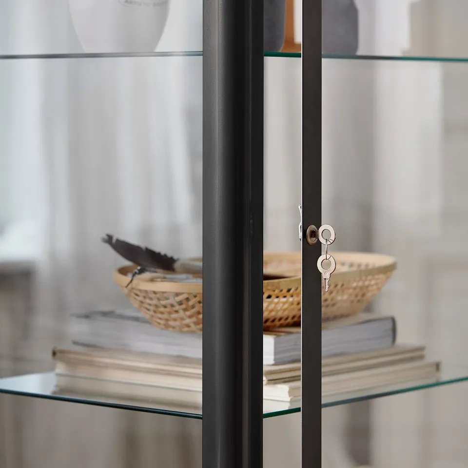 Glass-door cabinet with black metal frame displaying books and a basket with keys.