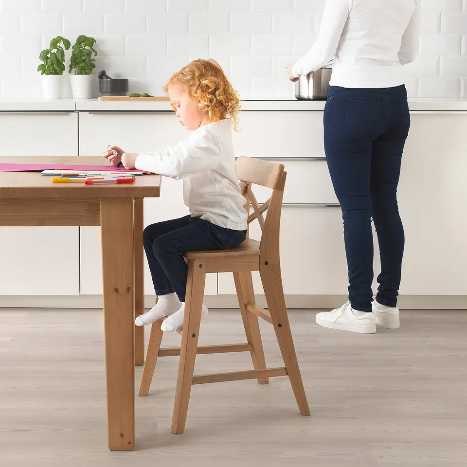 Child sits on a wooden junior chair at a table, drawing with crayons. Adult stands nearby.