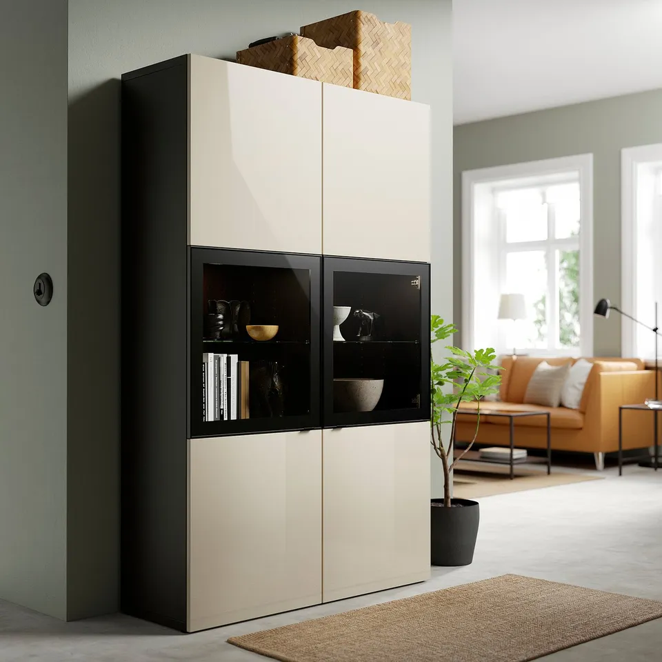 Modern beige BESTÅ cabinet with black glass doors. Holds books and decor, beside orange couch and lamp.