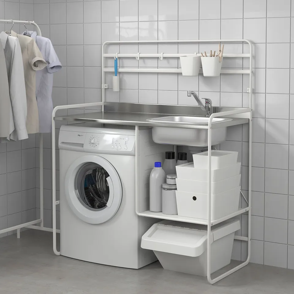 A compact, white SUNNERSTA mini-kitchen a sink, shelves, and storage bins, neatly organised beside a washing machine against tiled walls.