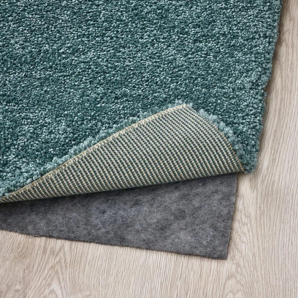 Green area rug corner with exposed gripper backing and rug pad on wooden floor