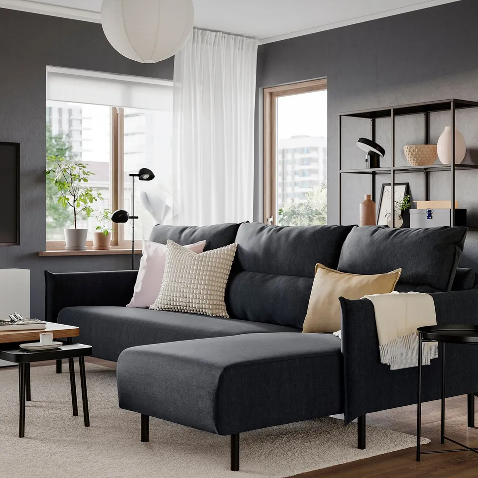 Dark grey sofa with chaise, plush cushions, by window, decorative pillows, throw blanket, side table with lamp & plant, nearby bookshelf.
