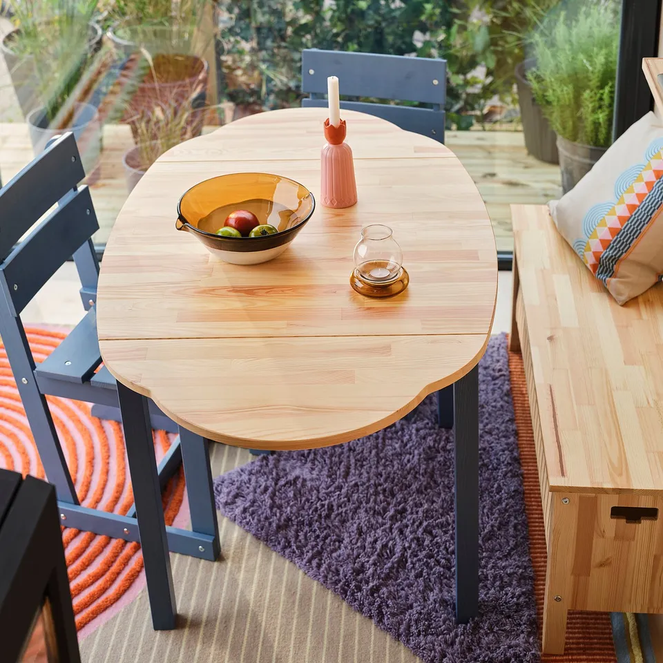 Blue-legged pine table with foldable leaves on purple rug. Holds fruits, vase, glass, with nearby bench.