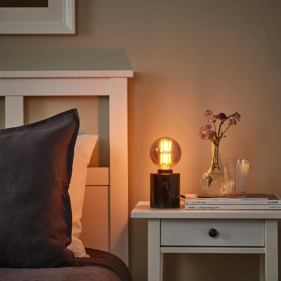 Cosy bedroom with MARKFROST lamp on bedside table. Black marble base, smoke glass shade, warm light.