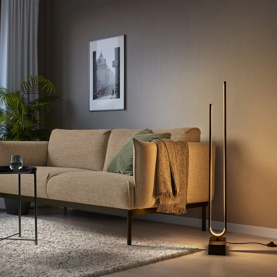 Contemporary living room with beige sofa, modern PILSKOTT floor lamp providing dynamic warm light, beside a small table with a drink and plant.