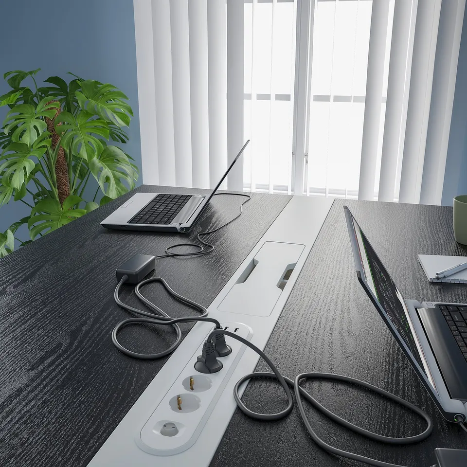 Modern high conference table with integrated power outlets and cable management for sitting or standing. A potted plant adds greenery beside windows.