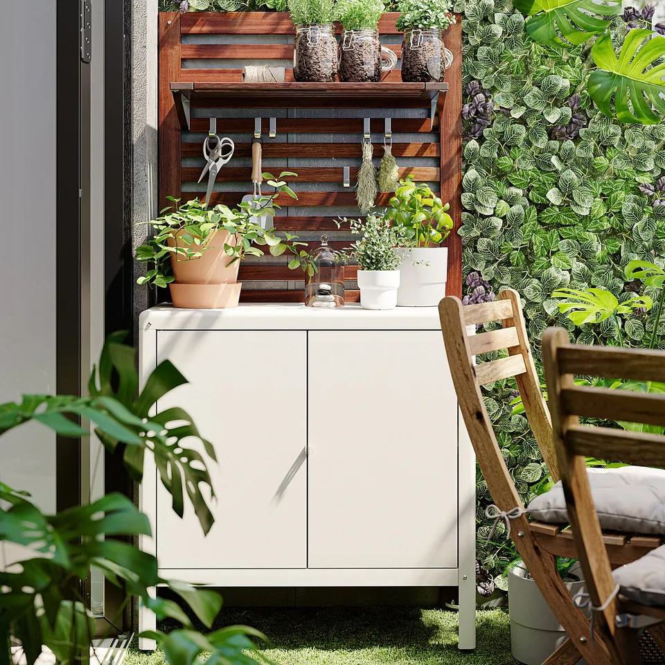Small white KOLBJÖRN cabinet indoors on a patio, surrounded by green plants and a wooden chair.