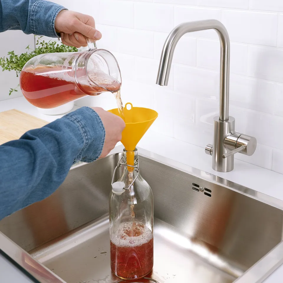 Person pours red liquid from pitcher into a clear bottle using a bright yellow funnel in a stainless steel kitchen sink.