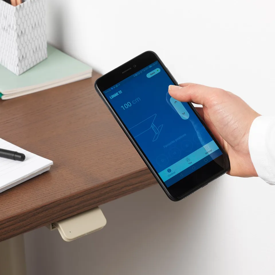 Person adjusting height of IDÅSEN desk using smartphone app. Desk is wooden with cable management underneath.