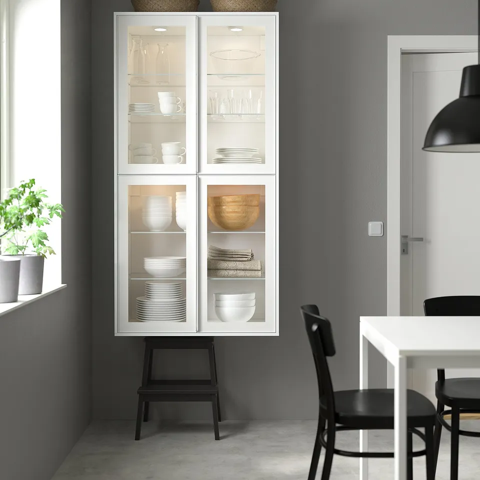 White IKEA HEJSTA glass cabinet against grey wall. Houses dishware, folds, and trays. Black base, pendant lamp, and chairs.