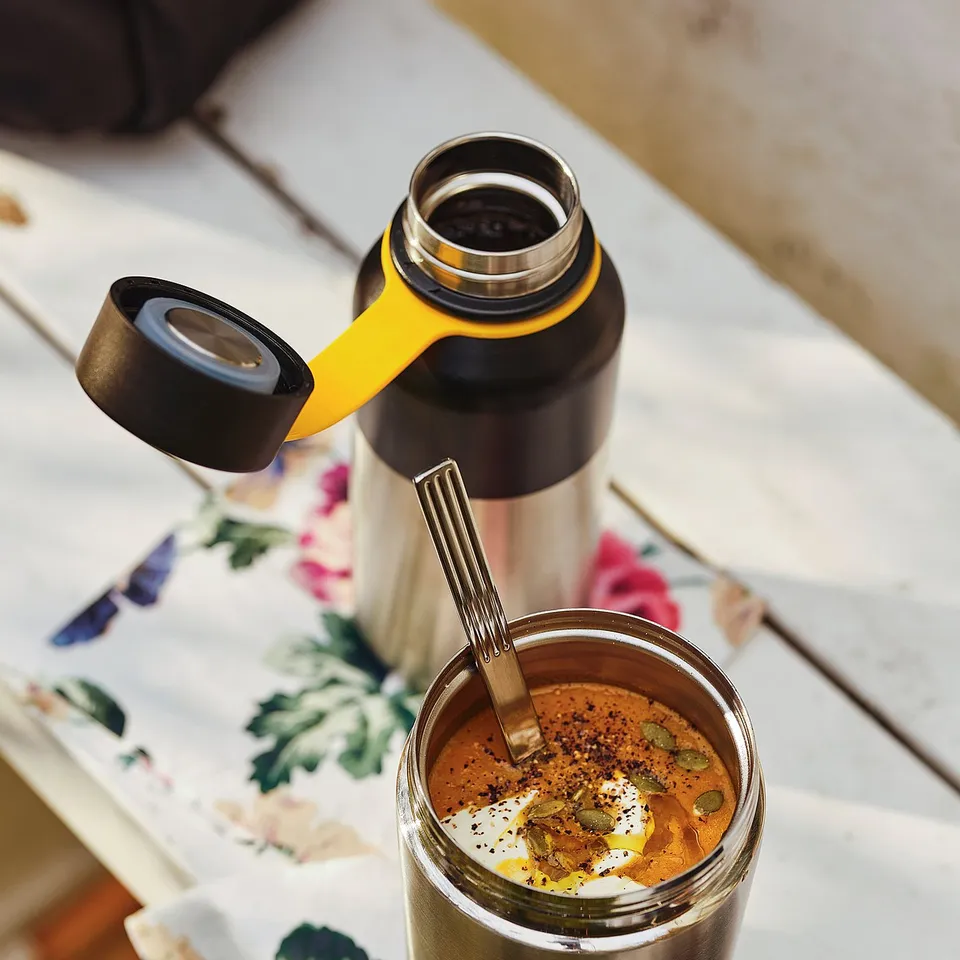 A thermos filled with soup sits on a floral cloth, accompanied by a bottle with a yellow handle.