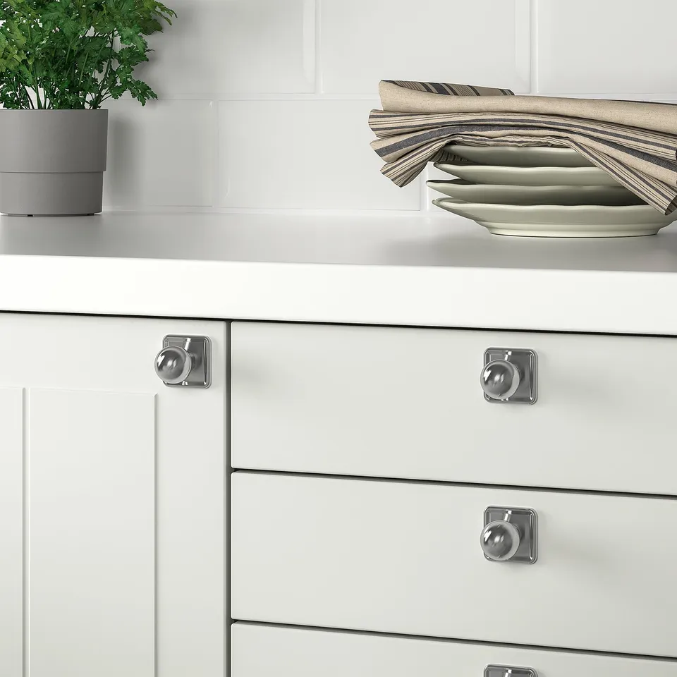 White kitchen drawers with silver knobs and handles, stainless steel plates, and a potted plant.