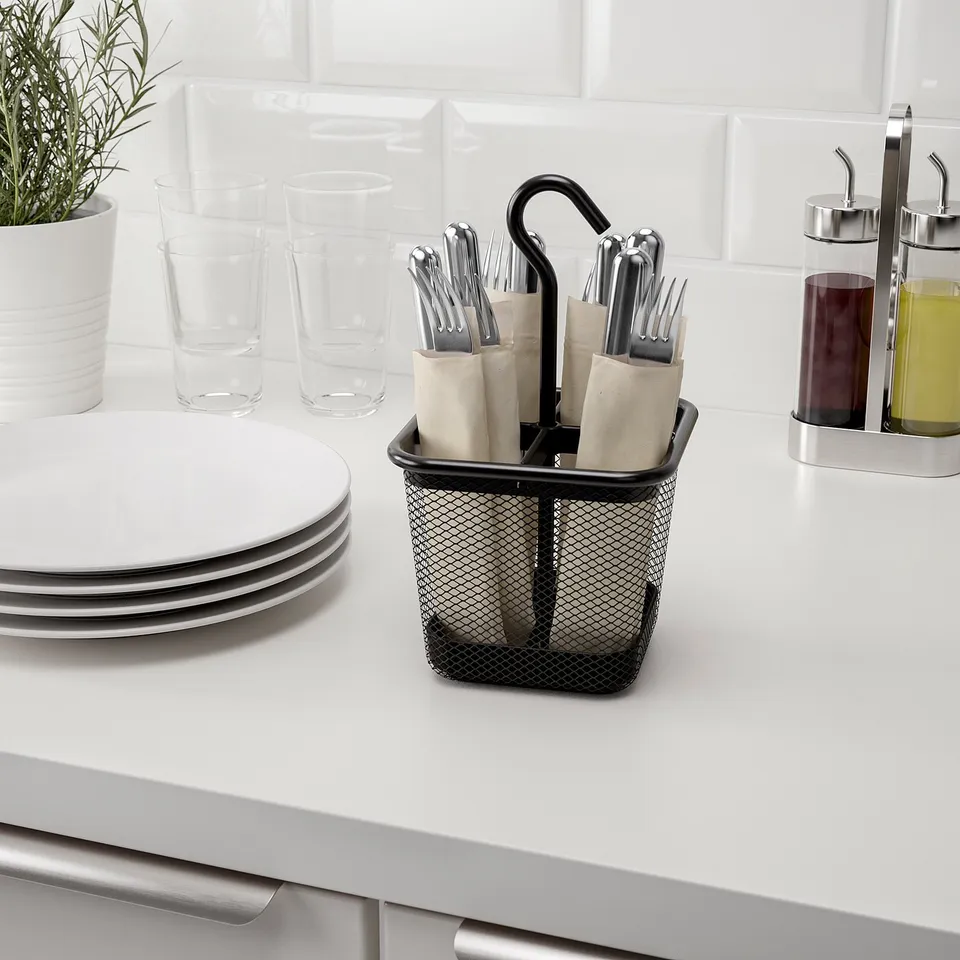 Black mesh cutlery holder with handle sits on counter, holding silverware with plates, glasses, and oil dispensers nearby.