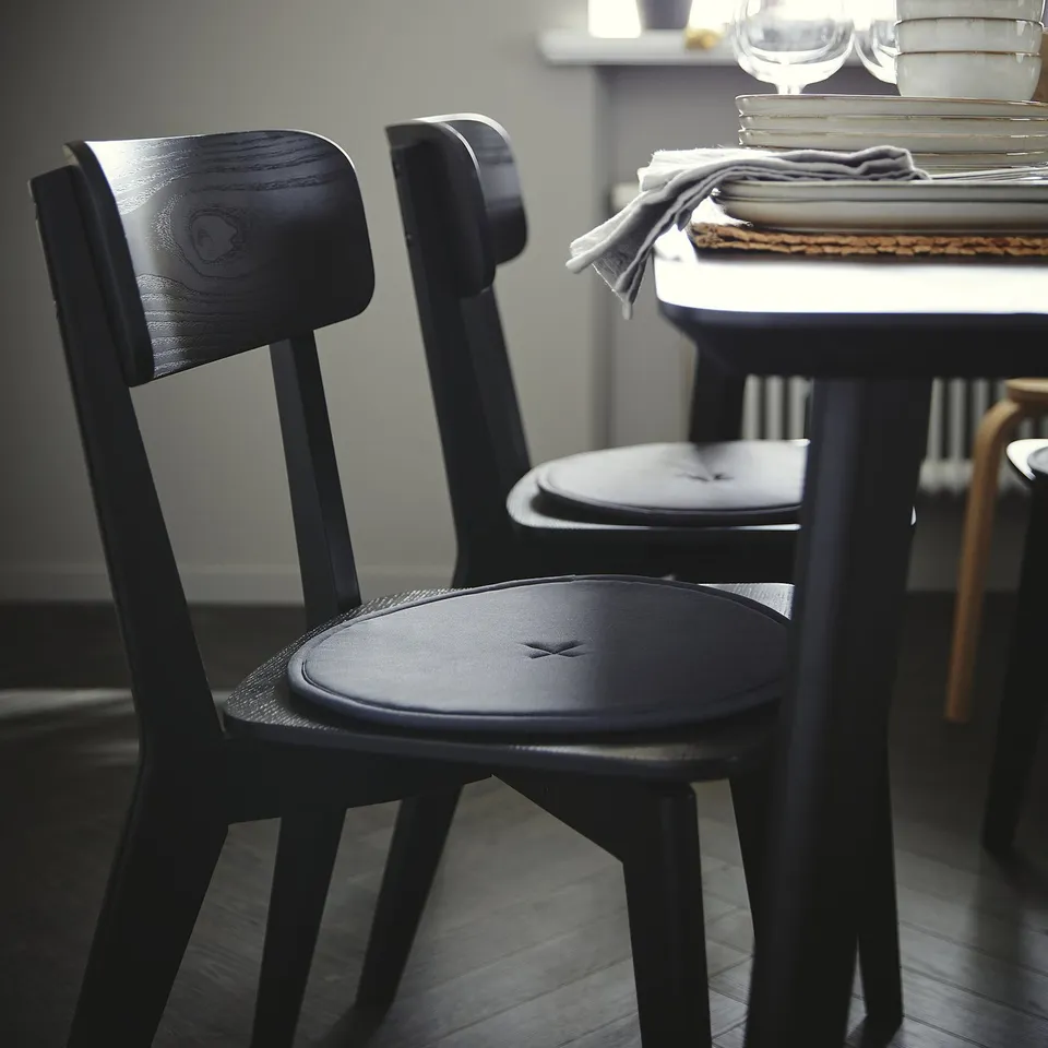 Two dark wood LISABO chairs near a table with stacks of plates and glasses. Minimalist design, sturdy, and compact.