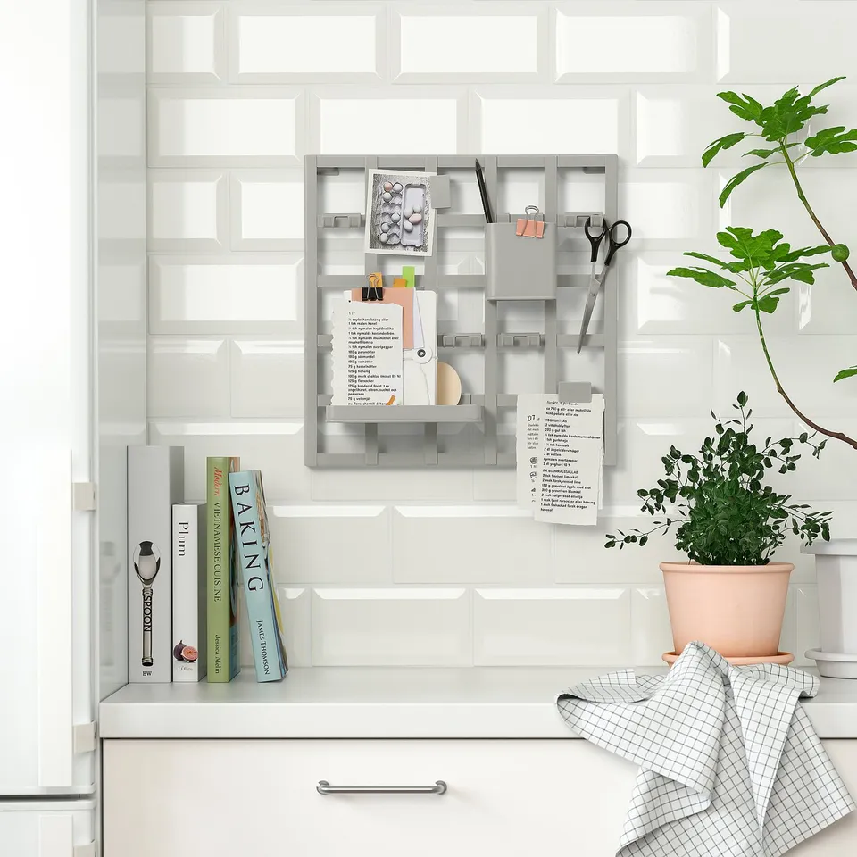 Kitchen wall organiser holds notes, scissors, books, with a shelf and hooks. Plants add greenery, towel draped.