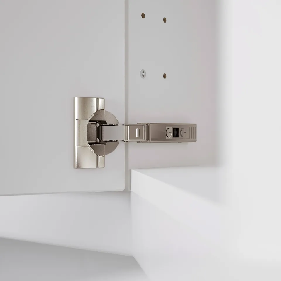 A modern kitchen hinge, adjustable in three directions, mounted on a white cabinet. Ideal for precise door alignment and easy removal for cleaning.