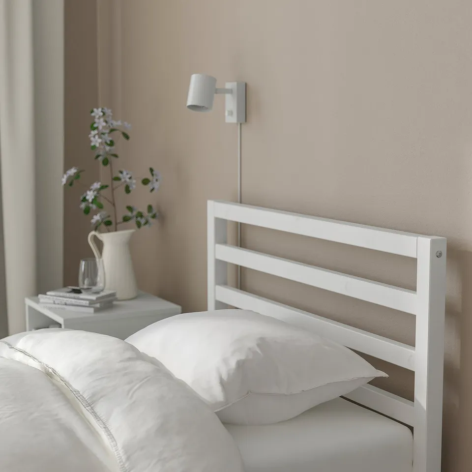 Bedroom: white bed, bedside table with flowers and books, wall light.