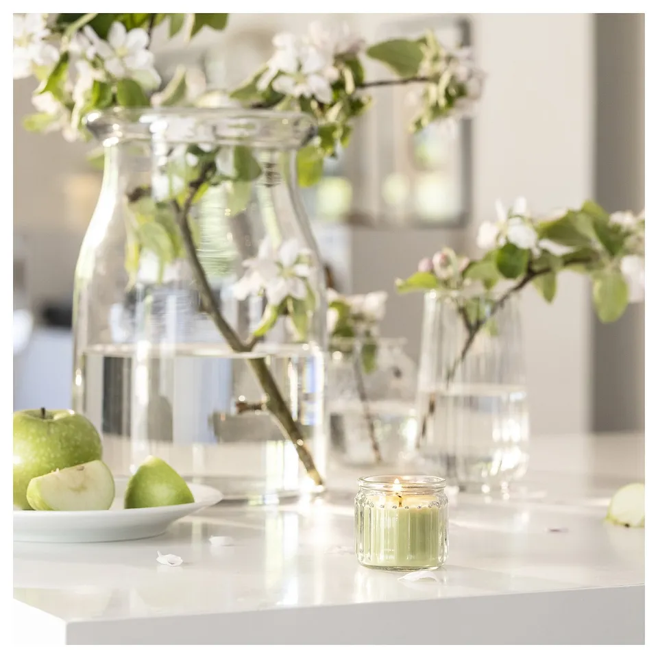 Clear glass candle, green wax, lit wick, apples, flowers in vases. Clean, fresh setting.