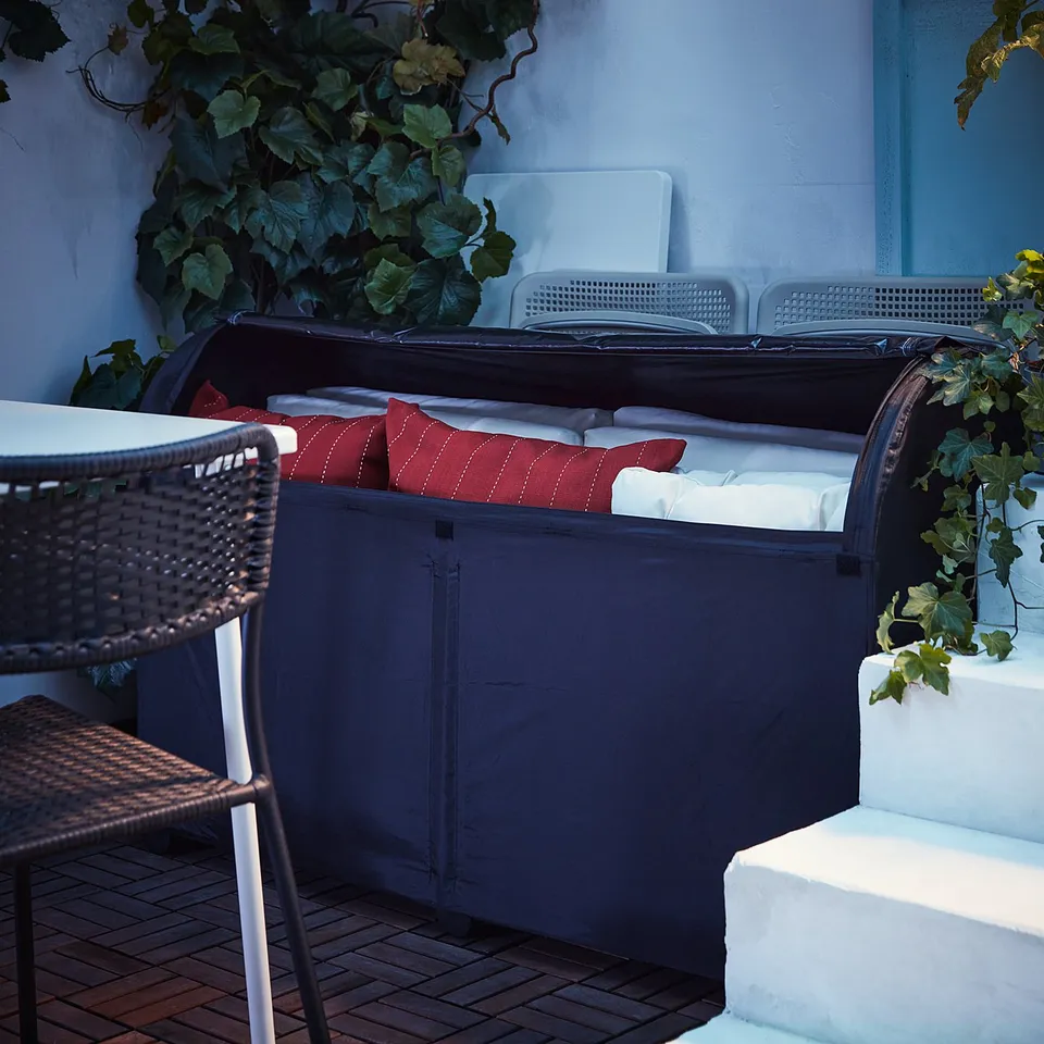 Dark grey outdoor storage box with red cushions, surrounded by green plants and white chairs.