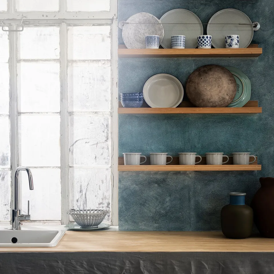 Kitchen sink under window with blue wall, bamboo shelves holding plates, bowls, mugs, and vases.