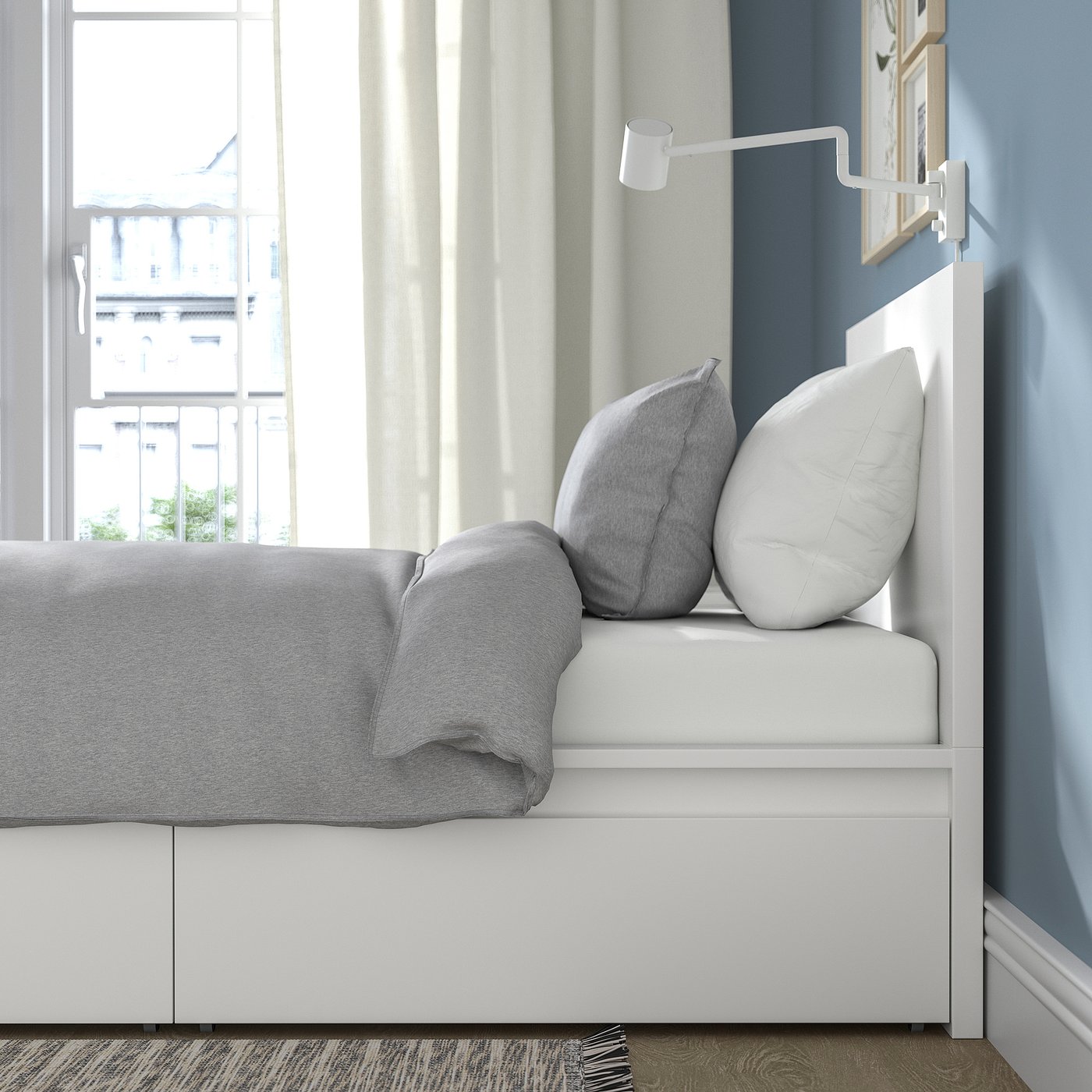 Modern bed with grey bedding, two pillows, and a white headboard against a blue wall. A window with curtains is visible in the background.