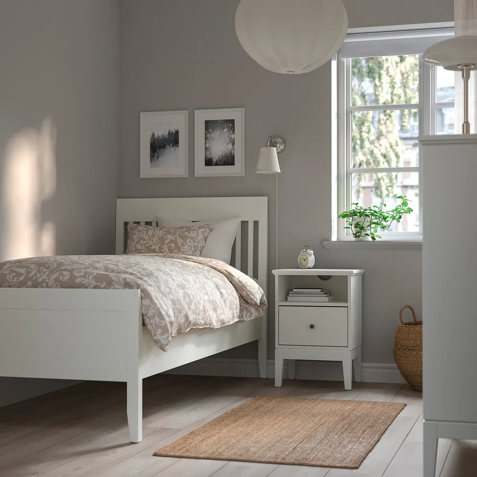 White IDANÄS bed frame with intricate headboard against purple wall, surrounded by bedside table with lamp, art, and plant.