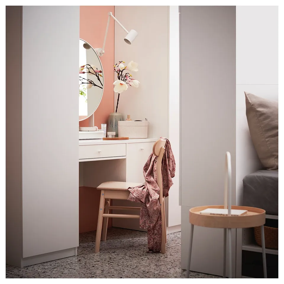 A minimalist white dressing table, SYVDE, with two drawers, glass top, and round mirror. Positioned against a light pink wall, adorned with flowers.
