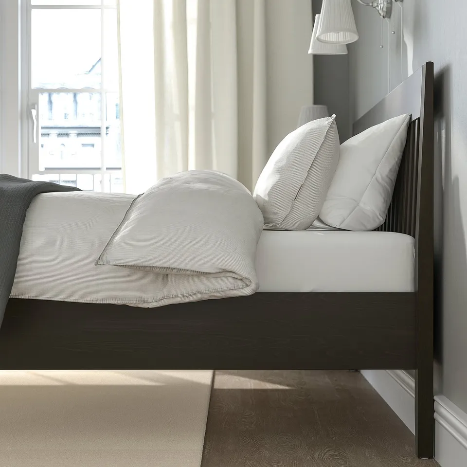 Modern bed frame with white bedding, large pillows, wooden headboard. Soft natural light from window.