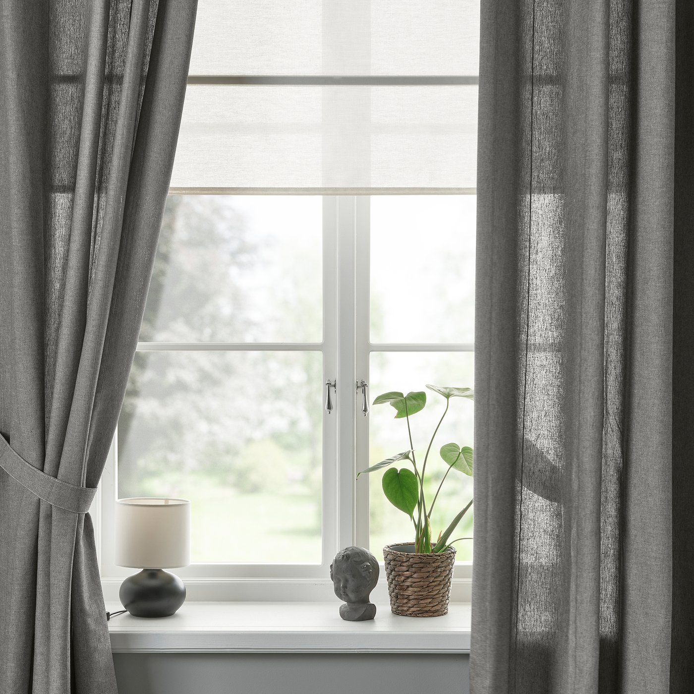 Window with grey curtains tied back, showing view outside. White sill holds lamp, plant, decoration. Light philtres through.