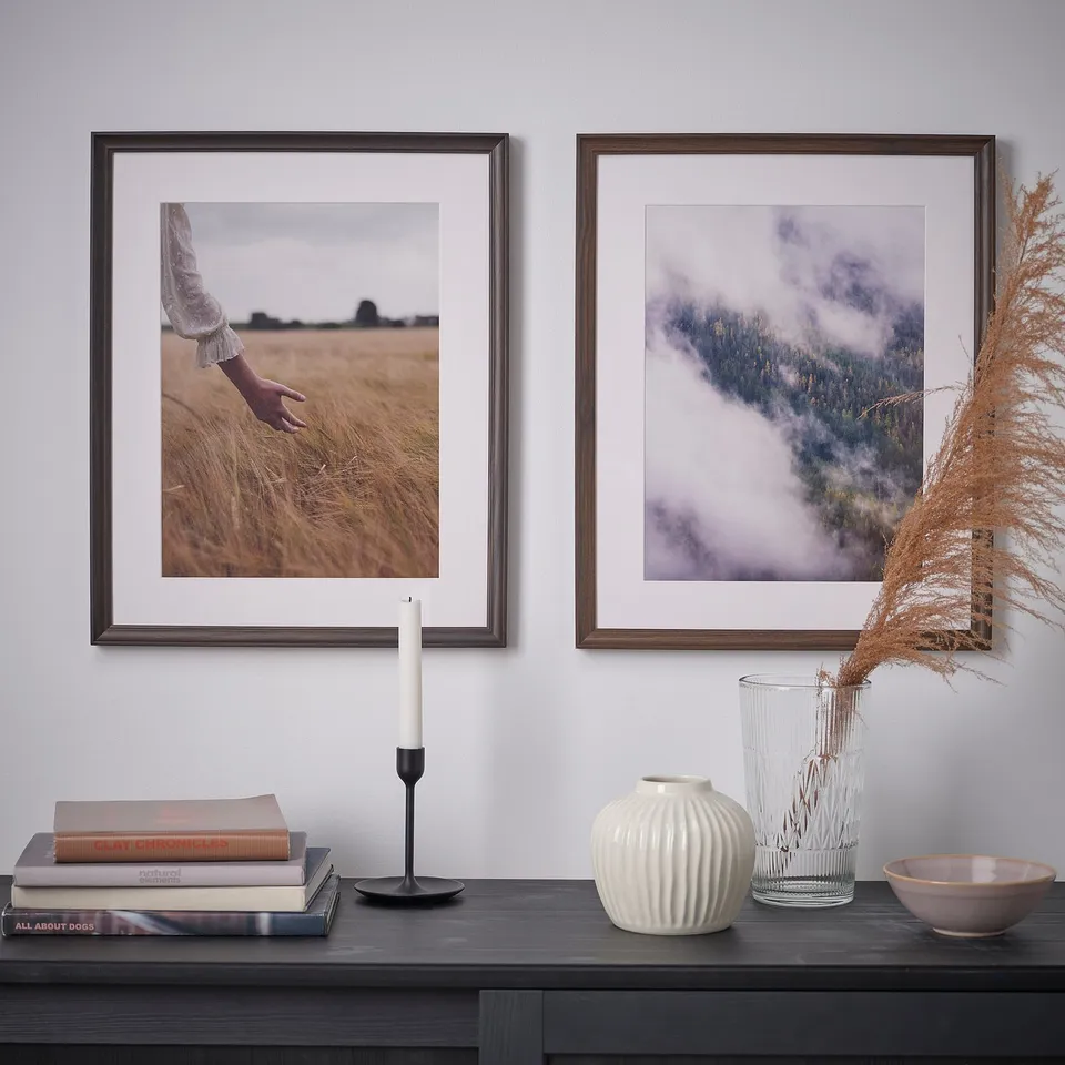 Two framed photos on wall: hand, grass; foggy landscape. Below, shelf with books, candle, vases, bowl.