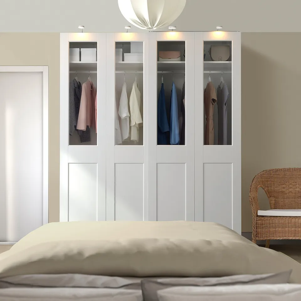 Modern white PAX wardrobe with glass doors clothes, lit from top. Also a wicker chair and large bed, emphasising its large size.