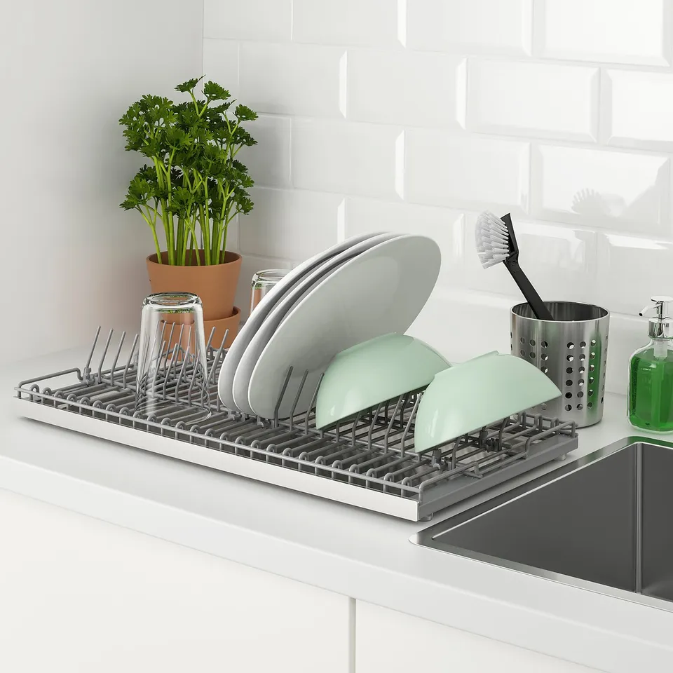 A stainless steel dish drainer on a counter, holding green and white bowls, plates, and glasses. A potted plant and cleaning tools nearby.