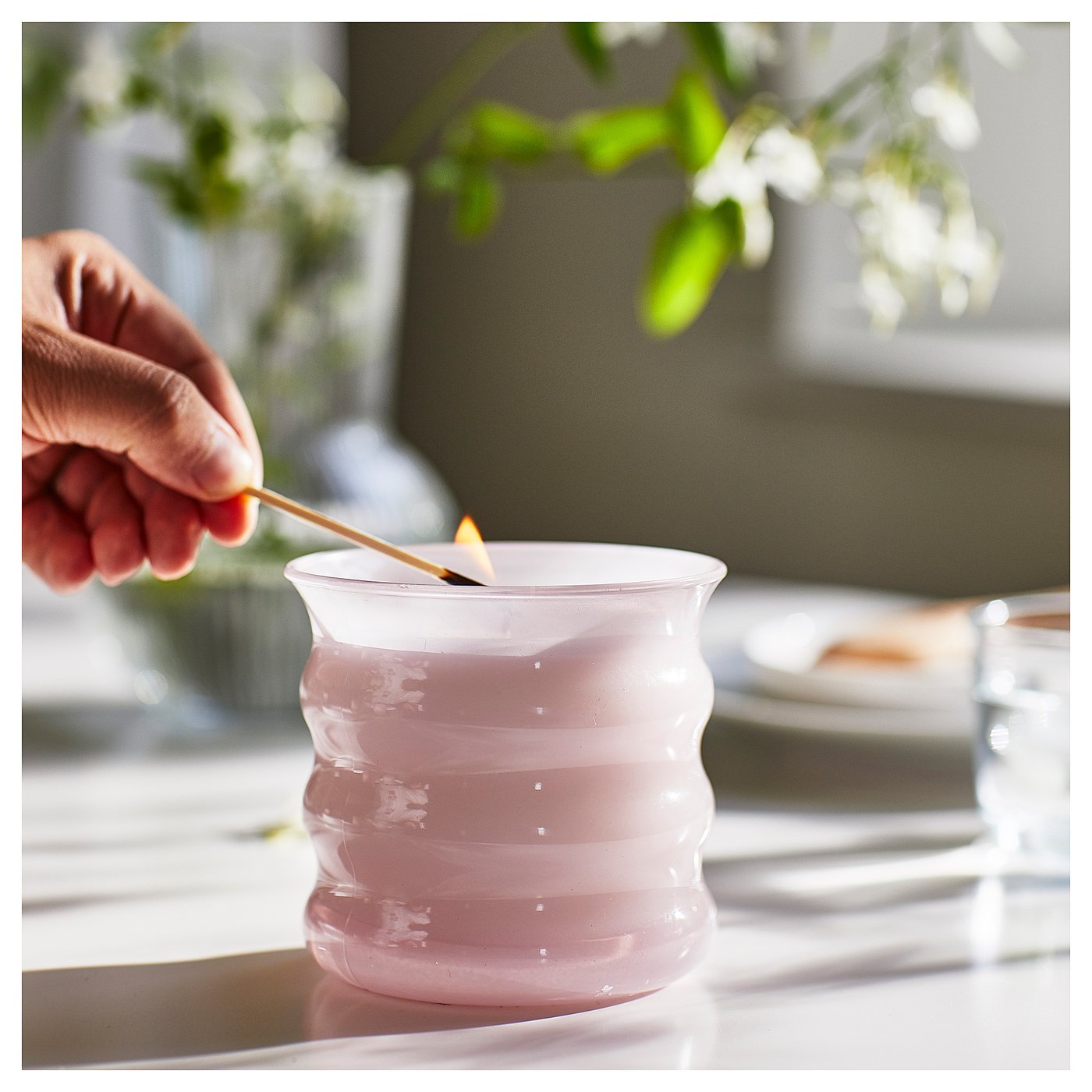 A hand stirs a pink LUGNARE scented candle in a clear glass holder on a table.