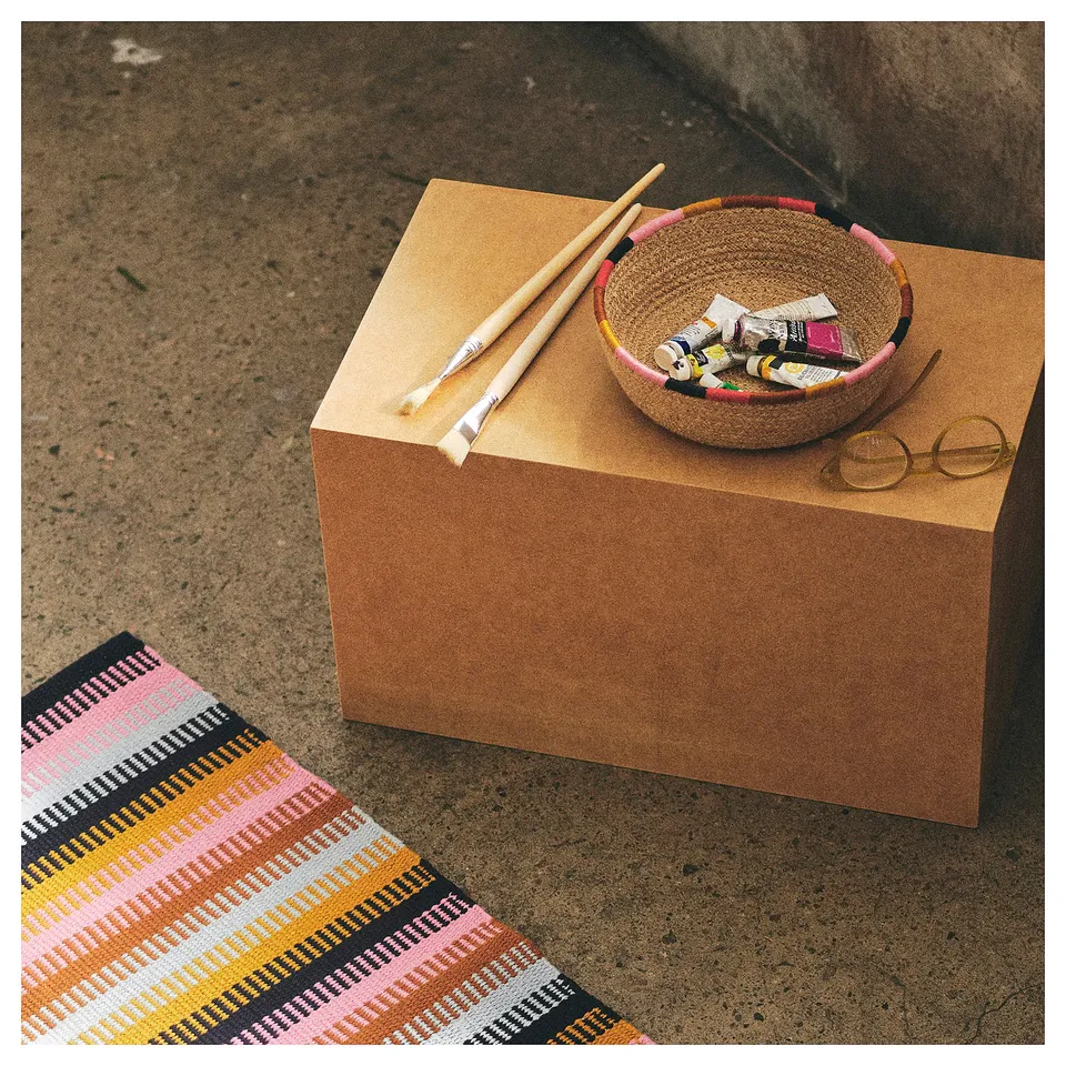 Jute MÄVINN basket with art supplies on cork cube, colourful rug, round glasses.