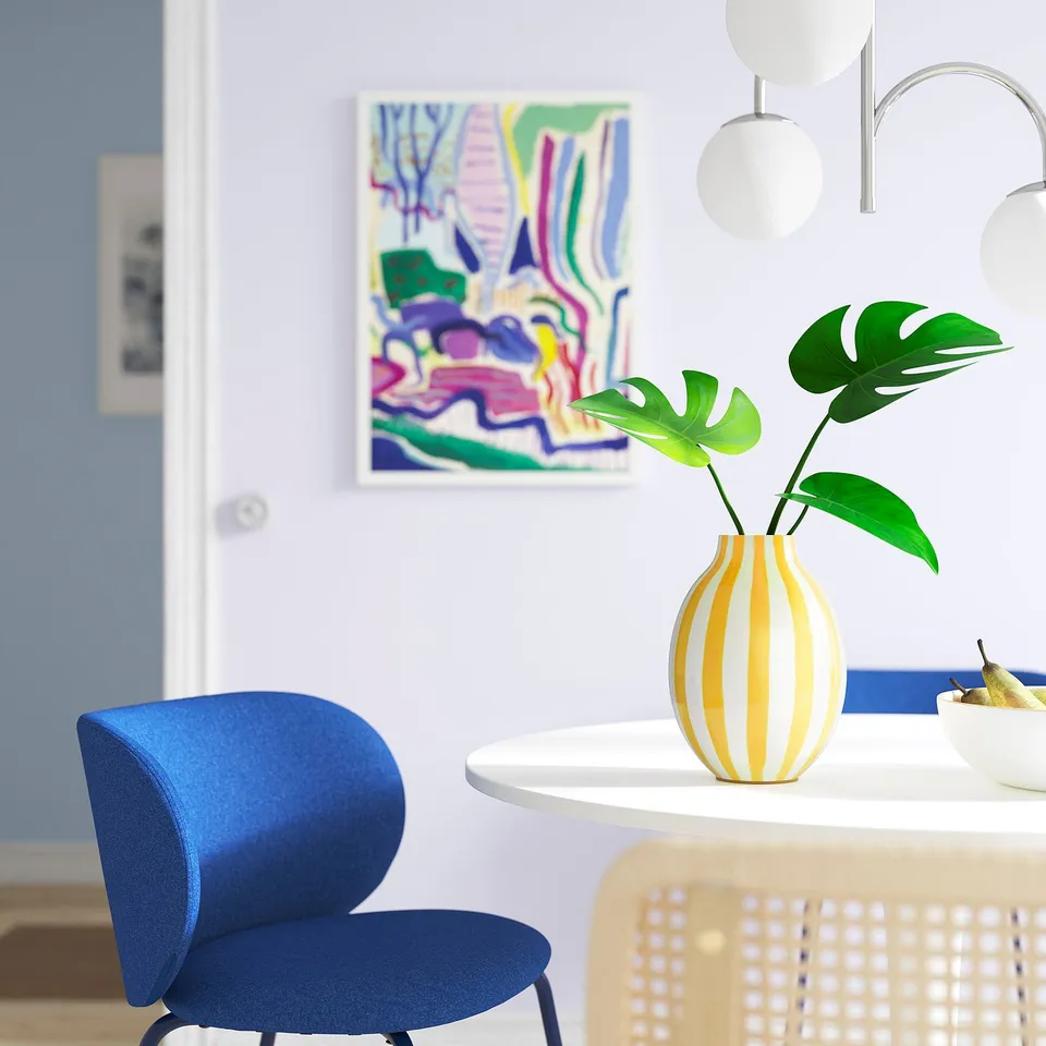 Bright yellow-striped BLODBJÖRK vase holds green leaves, sits on white table near blue chair. Modern, round, glossy stoneware.