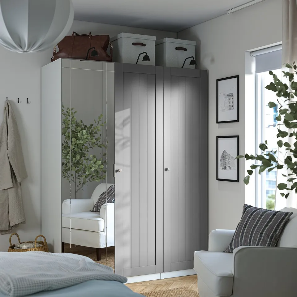 Modern bedroom with large grey PAX wardrobe, tall white chair, bed, and wall decor. Wardrobe is big, mirrored, with boxes on top.