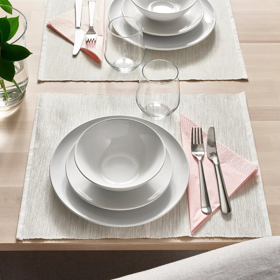 Elegant table setting with white plates, bowls, glasses, cutlery and napkins on woven place mats.