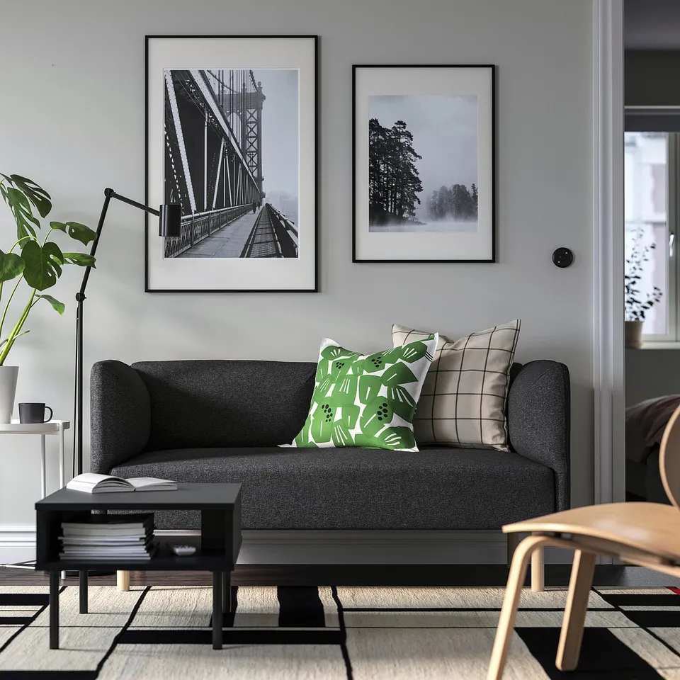 Modern living room with grey sofa, wooden chairs, green pillows, art, coffee table, books, mug, and plant.