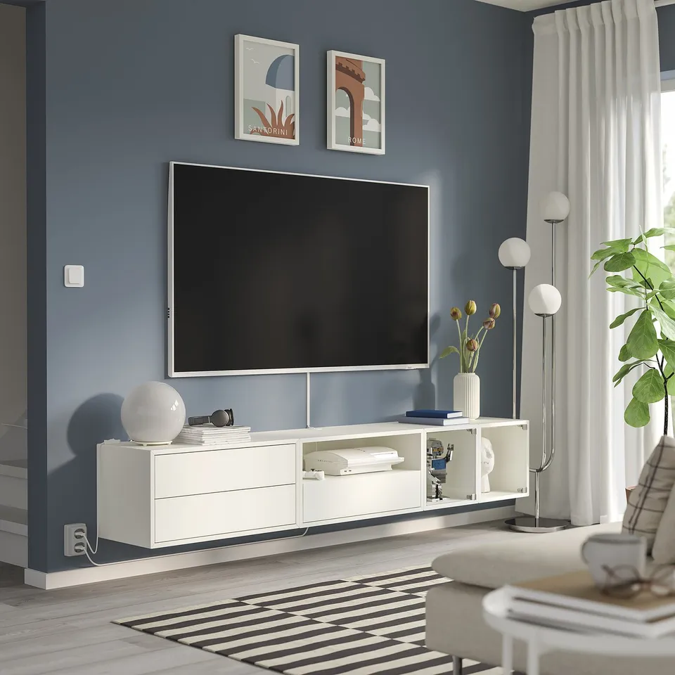 Modern living room: large flat-screen tv on white EKET cabinet, blue wall, striped rug, white lamps, plants, and artwork.