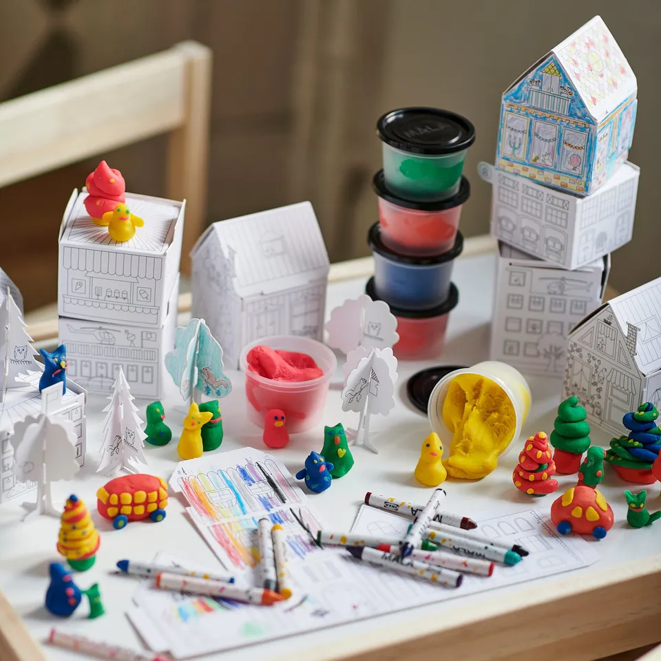 Kids craft table with MÅLA art supplies: colourful playdough, markers, and paper models of houses, trees, and cars.