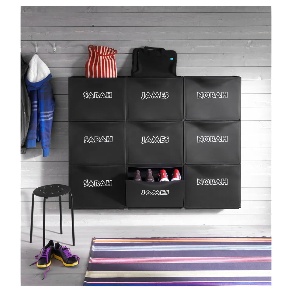 Black TRONES shoe storage units, personalised with names, against a wooden wall.