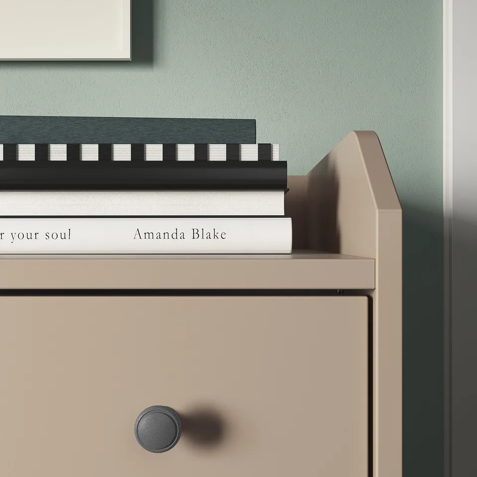 A HAUGA chest of drawers in beige with three drawers. The top holds a stack of books against a green wall.